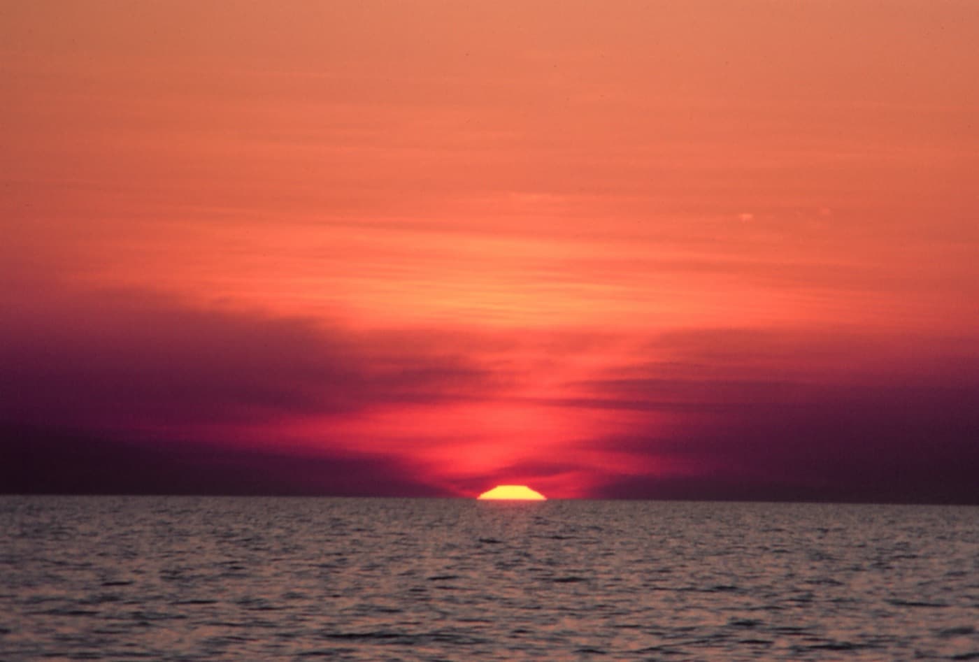 A photograph of the golden-red sun setting on the ocean horizon, its disc glowing low over the water beneath a sky of orange and crimson.