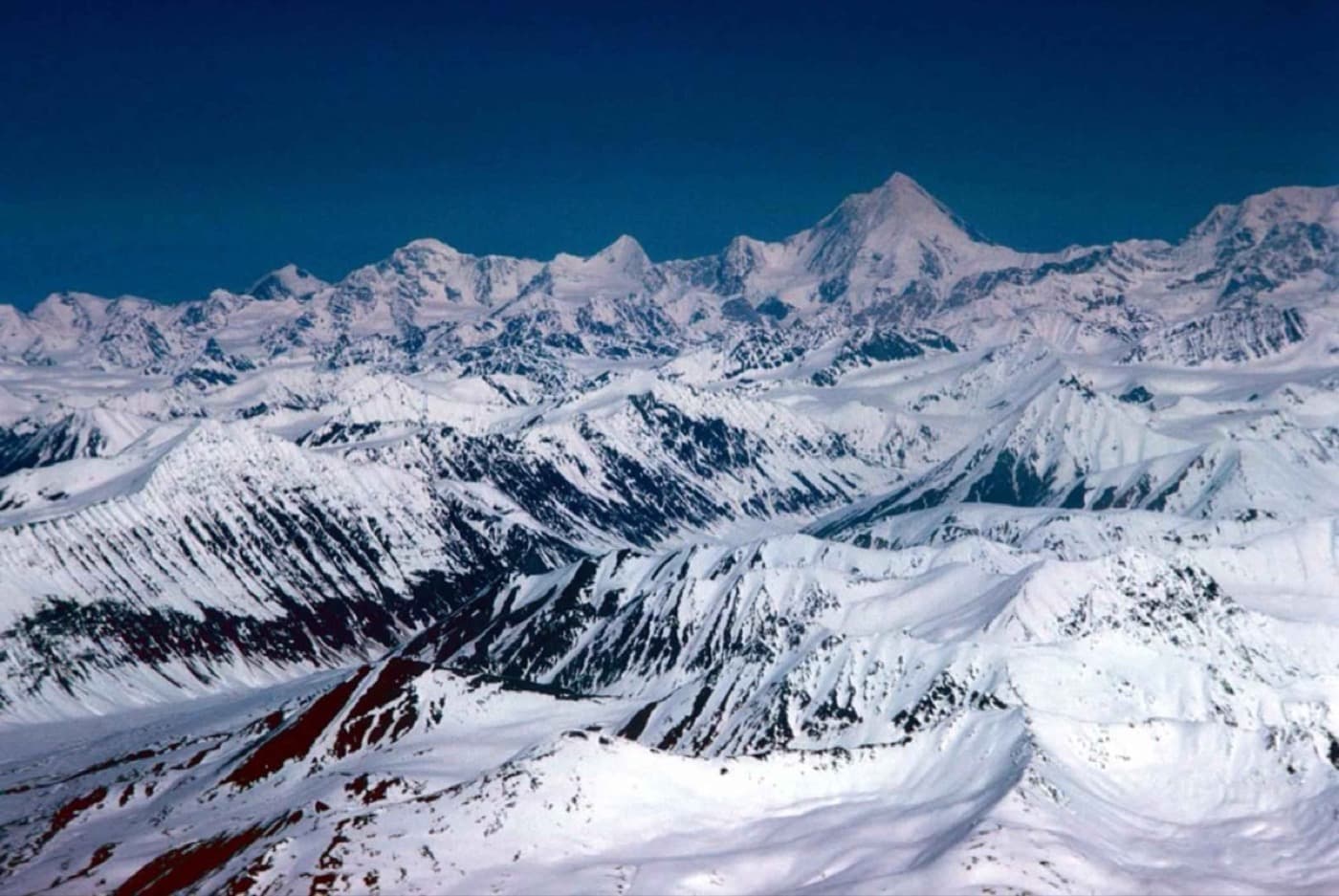 An aerial photograph of the snow-capped Alaska Range — sharp, rocky peaks rising in ridges above the surrounding land under a deep blue sky.