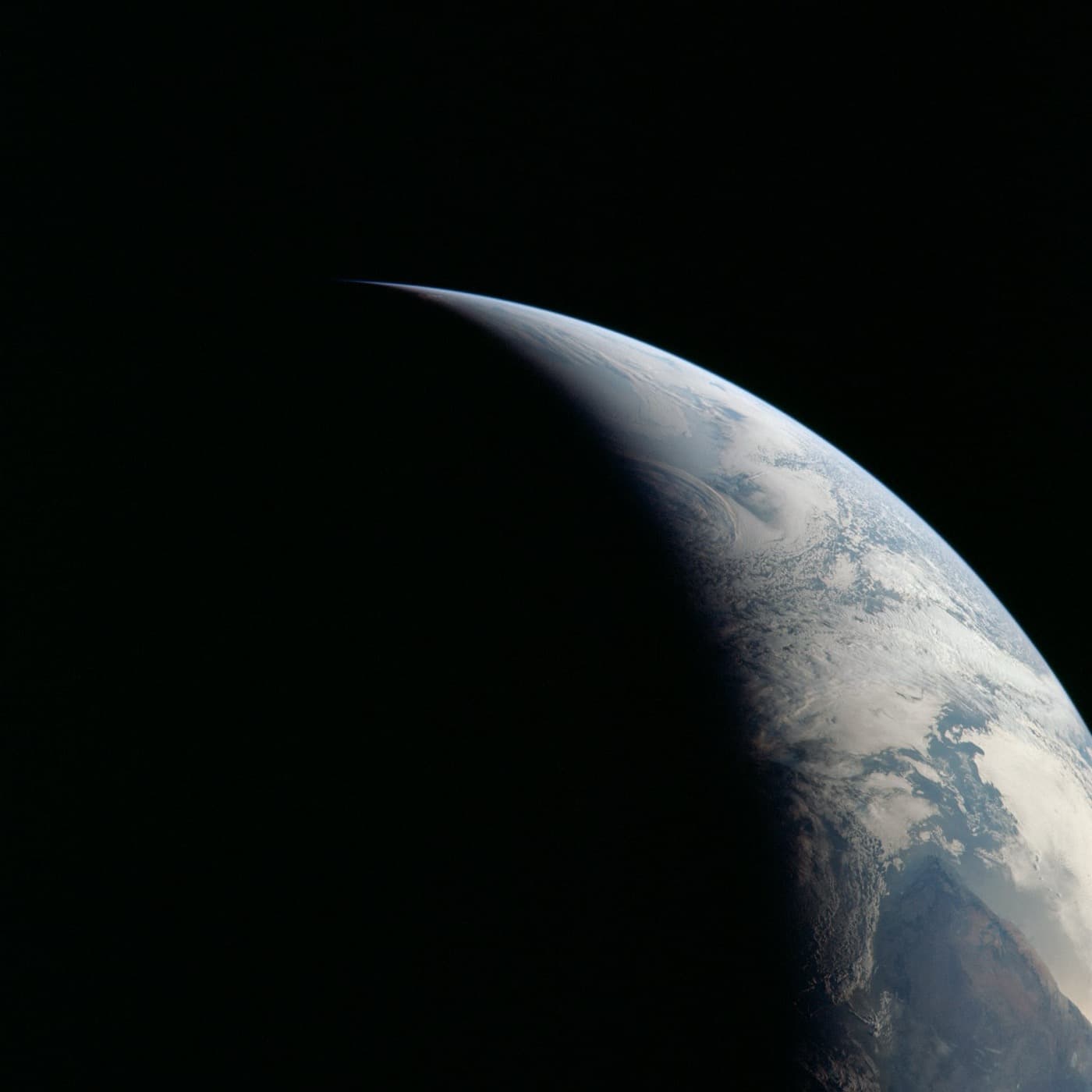 A photograph of Earth from space as a lit crescent: the sunward half glows blue and white in daylight while the curved terminator falls away into the dark of night.