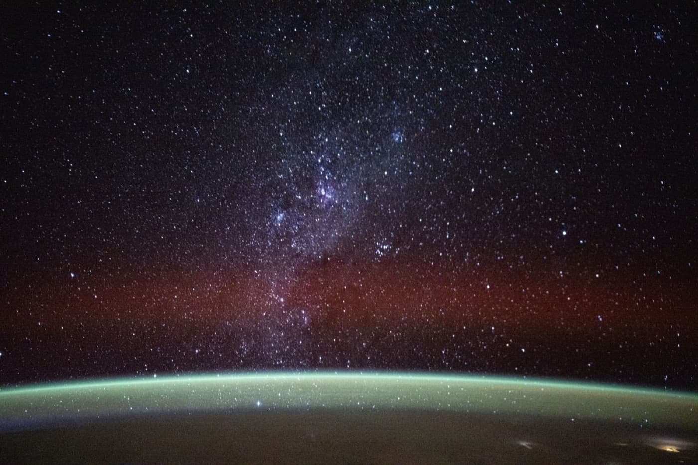 A long-exposure photograph of the night sky — a dense field of stars spread across the glowing band of the Milky Way, with the faint green airglow of Earth’s limb below.