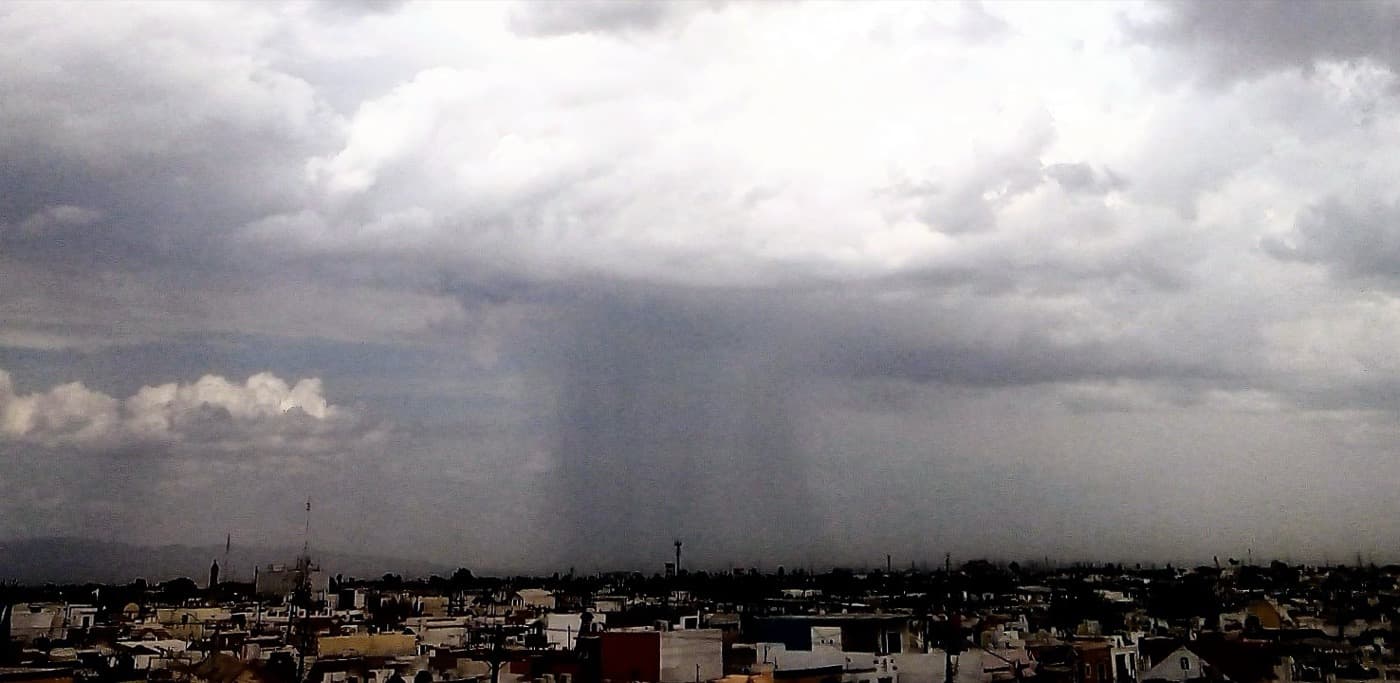 A photograph of a rain shaft — a dense vertical curtain of rain pouring from the base of a dark storm cloud down to the land below.